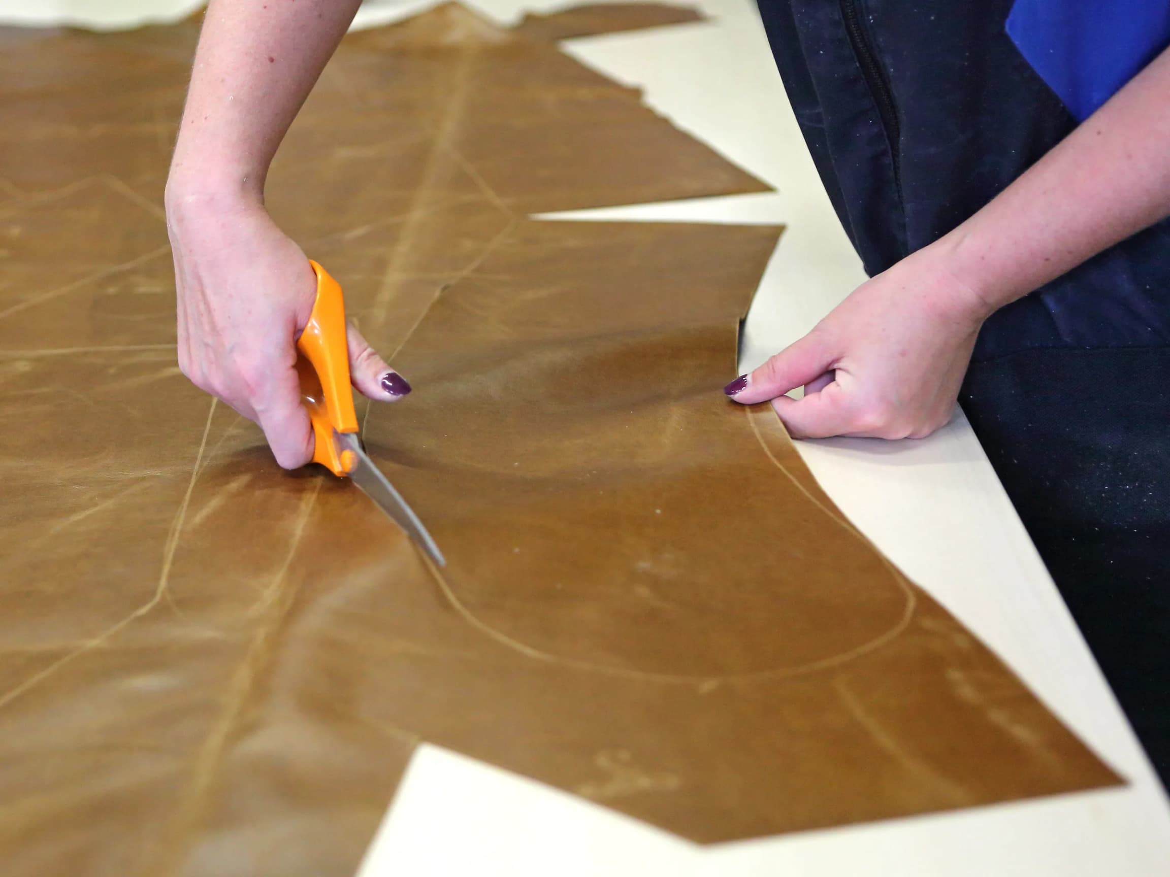 Hands cutting a leather hide along a marked pattern line with fabric shears, on a cutting table.